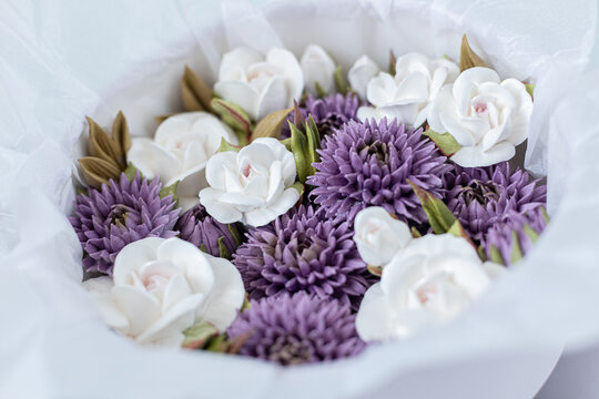 Floristry from Zephyr. White and purple artificial flowers arranged in a round bouquet with green leaves, wrapped in white tissue paper for decorative purposes