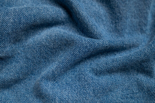 Blue denim fabric with soft waves fills frame in close-up. Denim texture and folded textile surface create versatile visual background for fashion, apparel, branding and design projects.