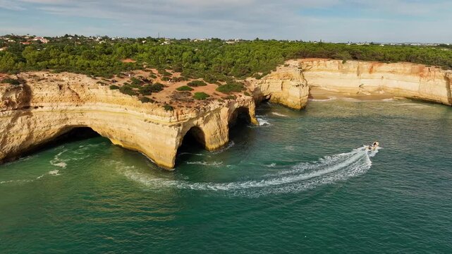 Drone view of Benagil Cave cliffs in Algarve, Portugal, with a tourist boat near limestone sea caves, turquoise Atlantic water, and rugged coast. Orbit parallax shot.