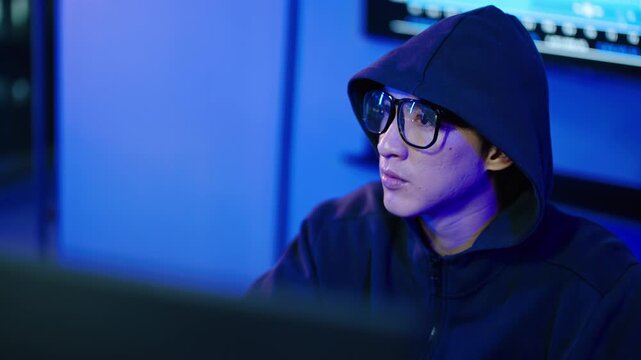 Focused Asian male hacker in a dark hoodie and glasses working on a computer late at night in a blue-lit room, analyzing secure cyber network data to execute a complex online digital system breach.
