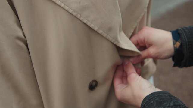 Close up of hands buttoning beige coat with dark fabric texture. action focuses on interaction between fingers and clothing emphasizing precise manual movement. Background remains