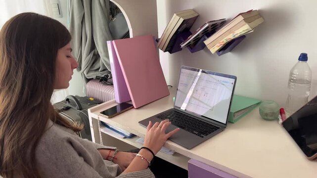 Teenage girl studying on a laptop at her desk in a bedroom, engaged in e-learning and online classes while attending remote education courses from home