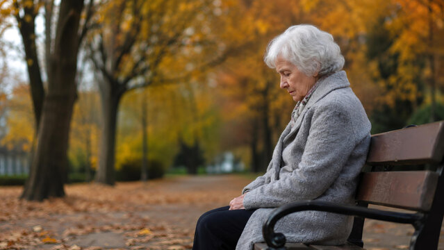 Sad senior woman sitting on park bench in autumn. Elderly female experiencing loneliness and mental confusion. Concept of aging, dementia, and emotional isolation in mental healthcare.