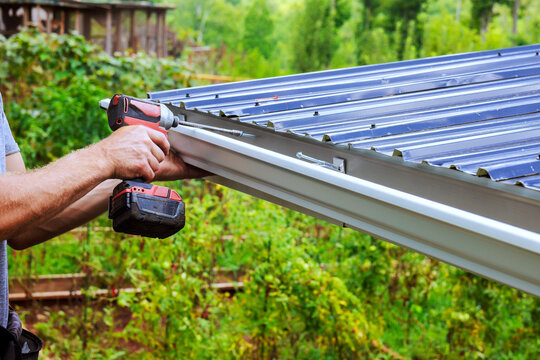 Contractor is attaching metal gutter sheets to roof using drill in garden.