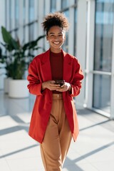 Obraz premium Happy professional woman in red coat walking with smartphone in sunlit office corridor for business banner and background design
