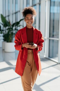 Happy professional woman in red coat walking with smartphone in sunlit office corridor for business banner and background design