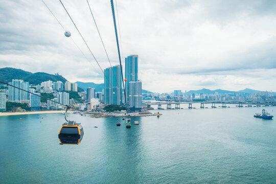 Cable car gondolas cross above the sea with Busan high-rise buildings skyline, bridge and mountains, South Korea. Elevated transport system, tourism infrastructure, coastal mobility, engineering