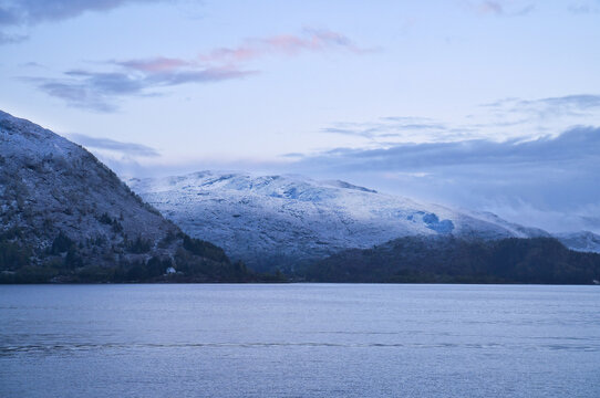 Snow-covered mountain range by a calm fjord at twilight