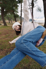 Naklejka premium Outdoor playful girl on a swing in a park, casual denim and sweater, laughing as she enjoys nature and leisure in a sunny autumn afternoon.