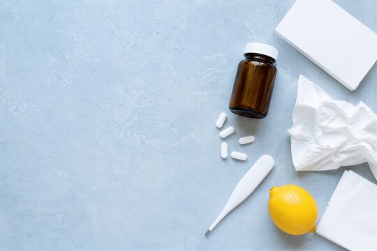 Cold and flu treatment concept with medicine bottle, pills, digital thermometer, crumpled tissue and lemon on blue background.