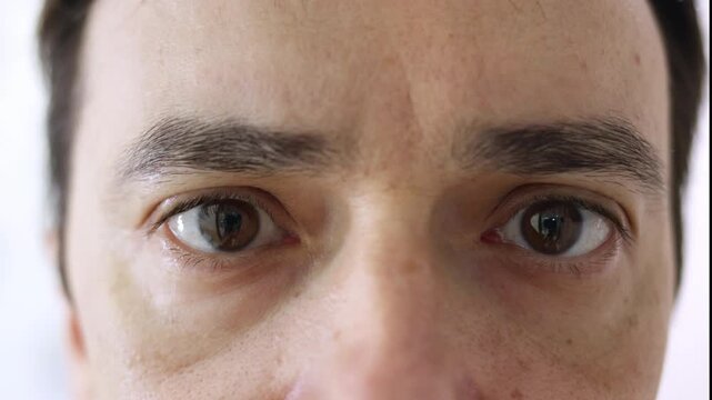Intense macro shot of a young man's brown eyes isolated on a white surface, staring directly into the camera while blinking slowly, showing every detail of the iris, pupil, and skin