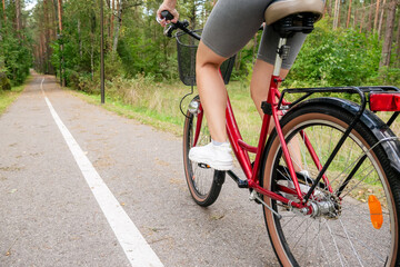Fototapeta premium Rear view of a young woman riding a red bicycle on a paved path stretching into a green pine forest. Active female lifestyle, outdoor cycling workout, summer fitness recreation, and eco-friendly city 