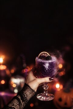 Woman hand holding glass of purple halloween cocktail with blackberry and dried orange slice. Spooky purple drink with smoke on dark background with candle light bokeh.