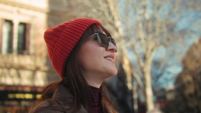 Profile portrait of a smiling young woman wearing sunglasses, standing on a city street with buildings in the background. Warm sunlight and first rays of spring create a soft, bright atmosphere. High