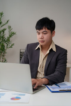 Young Asian businessman working on laptop, analyzing financial data and charts at office desk, managing business growth and planning corporate strategy for future success