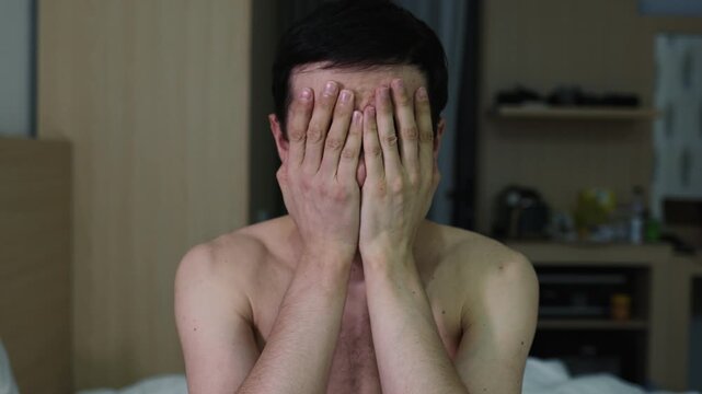 Upset and shirtless man sitting on a bed in his room, slowly covering his face with his hands in a gesture of despair, shame, or deep sadness, feeling miserable and emotionally overwhelmed
