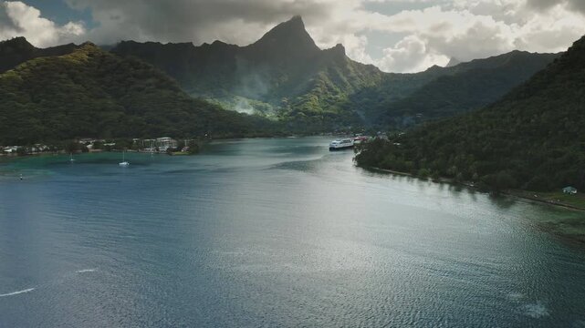 Moorea island Cook's Bay with its lush green mountains, Mount Rotui overlooking clear turquoise water, featuring ferry approaching the shore and sailboats anchored near the coastline. Aerial panorama