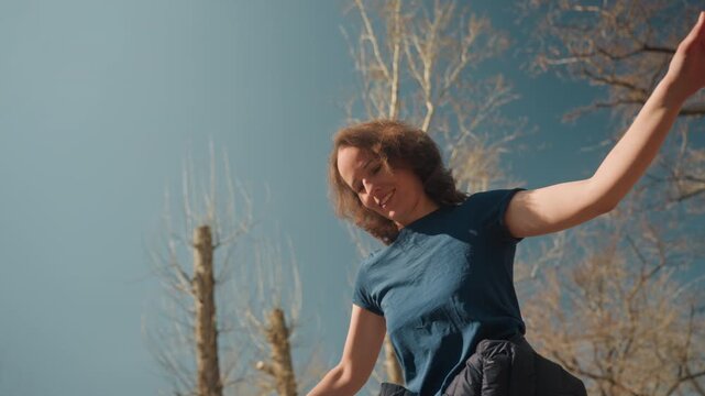 Close up of woman joyfully dancing outdoors on wooden structure with clear blue sky and bare trees in background. Her arms move freely as woman smiles wearing casual t shirt and carrying