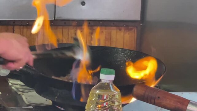 close-up kitchen motion of hand gripping wok while metal spatula stirs tossed food as large orange flames rise around black pan, clear plastic cooking oil bottle