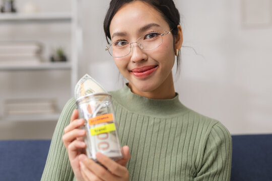 Travel savings and vacation budget concept with a young woman smiling and holding a money jar filled with cash at home, representing financial planning for a future trip and holiday.