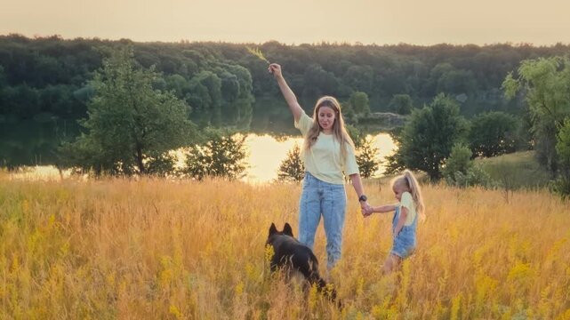 Happy mother and little daughter playing with their shepherd dog on a picturesque meadow near a river in Ukraine. Loving family spending time with their pet outdoors on a sunny day.