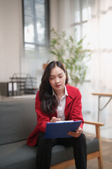 Fototapeta premium Asian professional woman sitting on a sofa, focusing on paperwork on a blue clipboard, embodying concepts of corporate work, business administration, and diligent analysis in an office setting