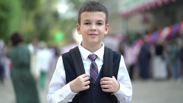 Portrait of smiling schoolboy in uniform and backpack standing near school with classmates and parents blurred in the background.