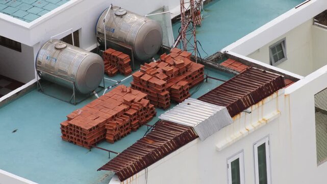 Elevated view of a building's rooftop in southeast asia, showing stainless steel water storage tanks, neatly stacked terracotta bricks for construction, and rusty corrugated iron sheets