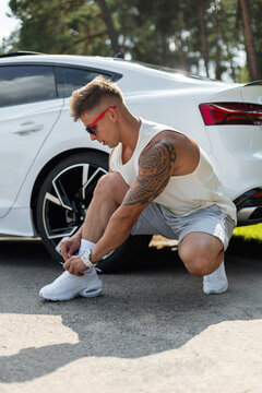 Muscular tattooed man crouching to tie his white sneakers next to a luxury white car on an outdoor road. Athletic guy preparing for a workout in sunny nature. Healthy lifestyle concept.