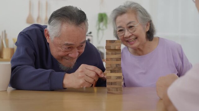 Asian senior man with friends playing wooden block tower game with concentration during indoor leisure activity. Active retirement lifestyle and wellness concept	