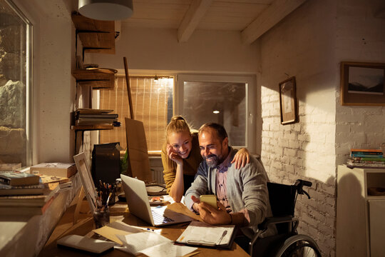 Wheelchair user and woman using laptop and smartphone at home office at night