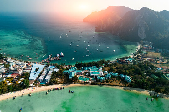 Resort hotels sit along the sandy beach and clear blue ocean water while numerous small boats float in the tropical bay under the warm light of the setting sun