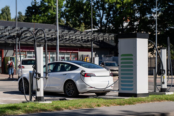 A white electric car charges at a public fast-charging station, the roadside infrastructure that decides how quickly a country can retire its petrol fleet. © K