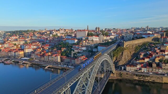 Drone view of Dom Luis I Bridge over Douro River in Porto, Portugal, with waterfront lights, including a tram on the bridge and rabelo boats by the quay at sunrise. Push in approach dolly shot