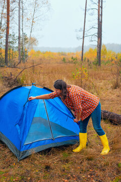 A woman in a plaid shirt and yellow boots Setting up a tenta blue tent amidst the dry grass and trees of a foggy woodland during fall
