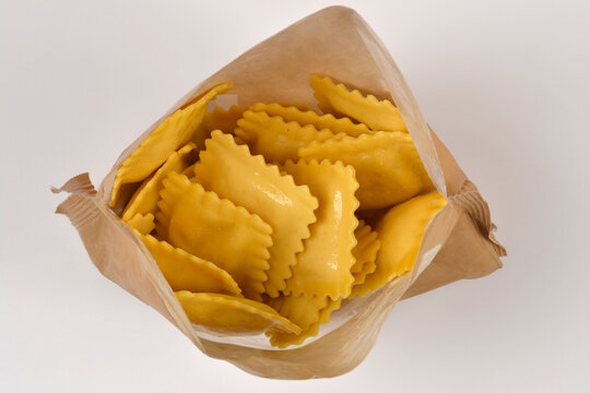 Top view of an open brown plastic pouch packaging filled with yellow raw uncooked square ravioli pasta on a solid white studio background.