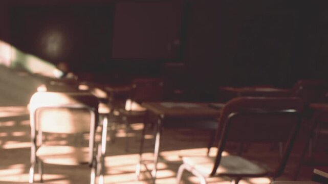 Desks and chairs are arranged in a classroom. Sunlight shines through windows, creating shadows on the floor. It is afternoon and the room is quiet.
