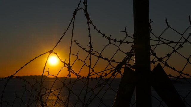 Barbed wire fence blocking setting sun over ocean. Golden sun setting over a tranquil ocean, partially obscured by the silhouette of a barbed wire fence, creating a contrast nature and limitation