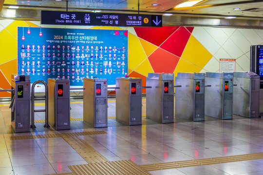 Metal fare gates, tactile paving, multilingual directional signs, and a large promotional display fill a colorful subway station interior.