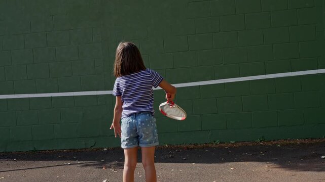 Rear view child practicing tennis outdoors against dark green cinderblock wall with white target line, racket follow-through, ball in play, casual sports training, urban court texture, sunlight