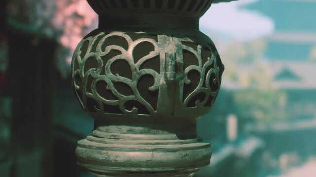 A metal lantern with intricate patterns stands in a garden. Soft light filters through the trees and highlights the craftsmanship. The setting suggests a quiet moment in a historical place.
