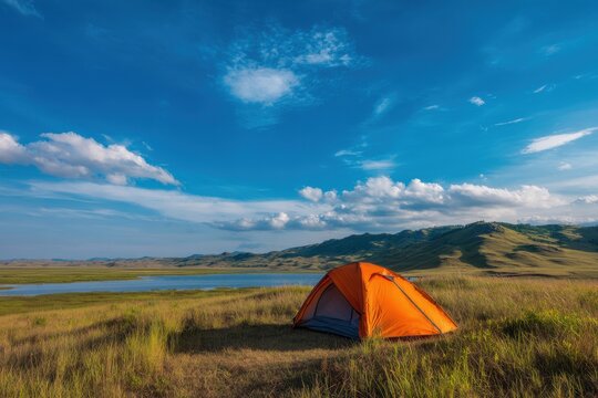 Orange camping tent pitched in a grassy field beside a tranquil lake under a clear blue sky with fluffy clouds adventure landscape vacation wilderness backpacker recreation basecamp