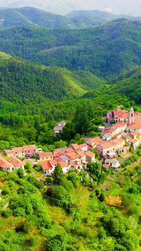 Vertical video. Carrodano Superiore, Italy. Medieval hilltop village with terracotta rooftops and a church tower, nestled in dense green forest of the Val di Vara, seen from above under soft daylight