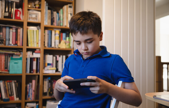 Focused Young Boy Using Mobile Phone While Sitting In Study Room, Child Holding Smartphone For Gaming And Online Learning At Home, Back To School, Digital Education, Technology And Screen Time.