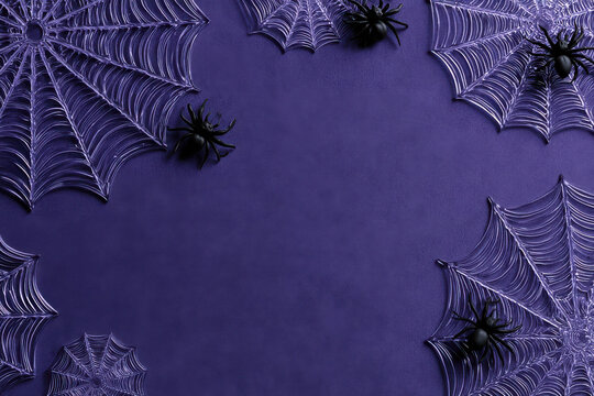 Purple halloween background with copy space, shiny plastic spider webs and fake black spiders on textured dark violet surface, flat lay frame arrangement