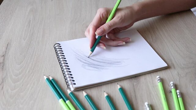 POV hand drawing with pencil in sketchbook on wooden table, artist hatching graphite sketch in spiral notepad with green pencils around, art therapy process, close up