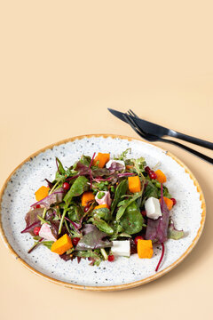 Plate with salad with greens, pumpkin and soft cheese and cutlery on beige background.