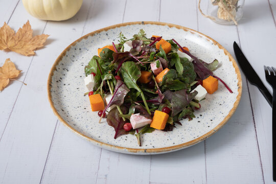 Plate with salad with greens, pumpkin and soft cheese and cutlery on a wooden background.