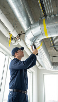 Caucasian man in work suit and glasses inspects metal HVAC ducts on ceiling using handheld device. A maintenance and safety check, building systems, tech, or industrial concept.