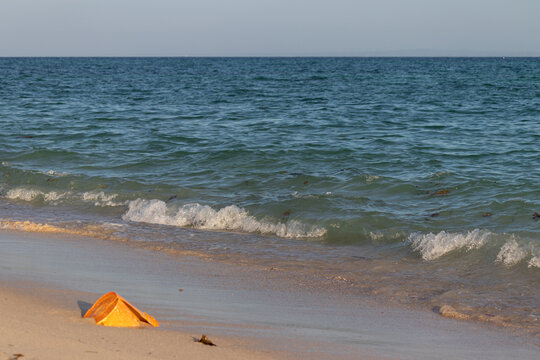 Broken orange plastic bucket on the sand, partially submerged in shallow water as gentle waves lap at its